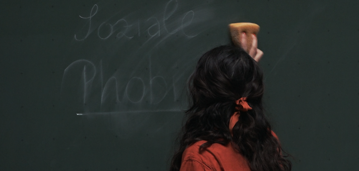 Junge Frau mit Schwamm vor einer Tafel. Sie wischt auf der Tafel die Worte "Soziale Phobie" weg.