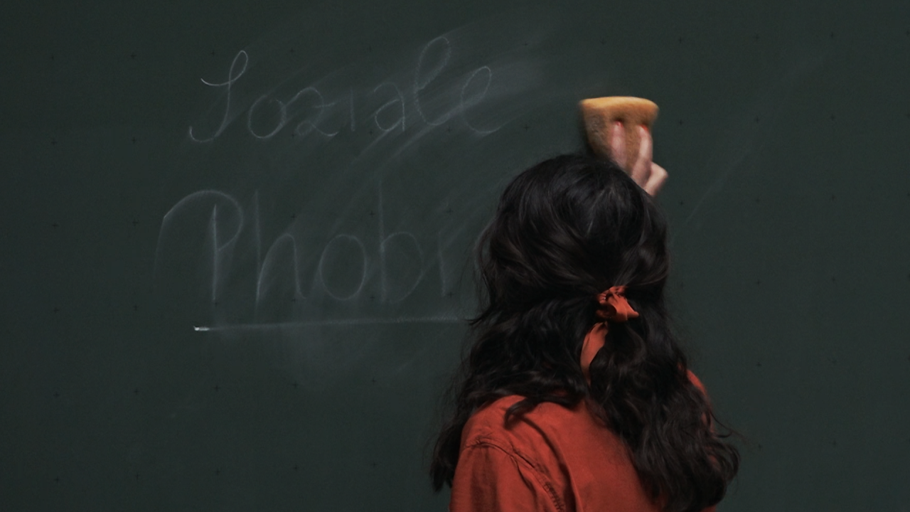 Junge Frau mit Schwamm vor einer Tafel. Sie wischt auf der Tafel die Worte "Soziale Phobie" weg.