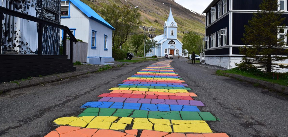 Ein Regenbogenweg in einer Kleinstadt der zu einer kleinen Kapelle aus Holz führt.