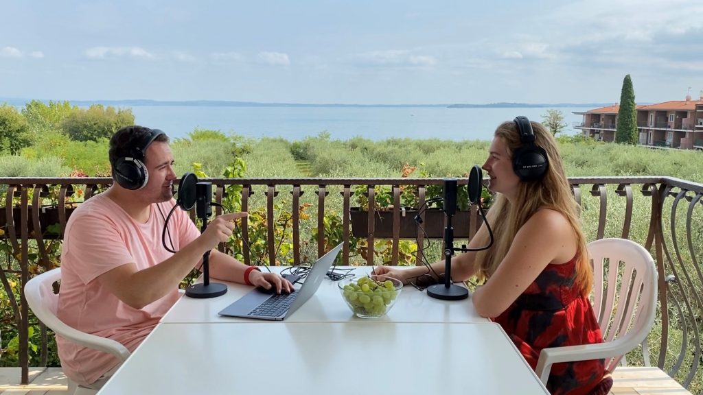 M94.5 Moderator:innen Simon Fischer und Nicole Albrecht in ihrem Freiluftstudio, im Hintergrund der Gardesee und Olivenbäume