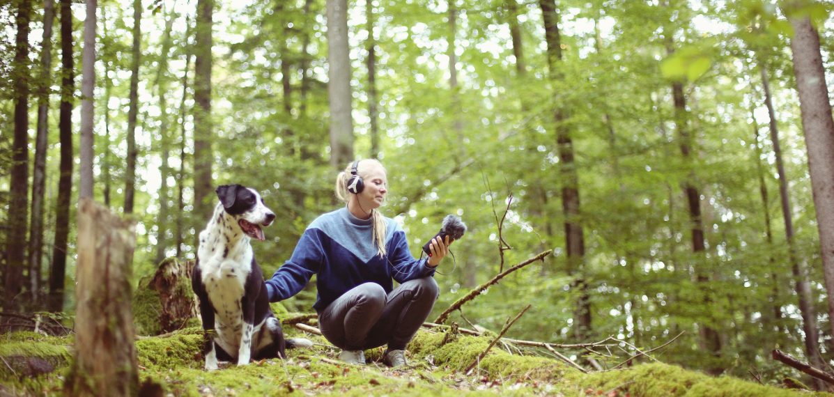 Frau (her tree) nimmt im Wald Geräusche auf. Neben ihr ein Hund.