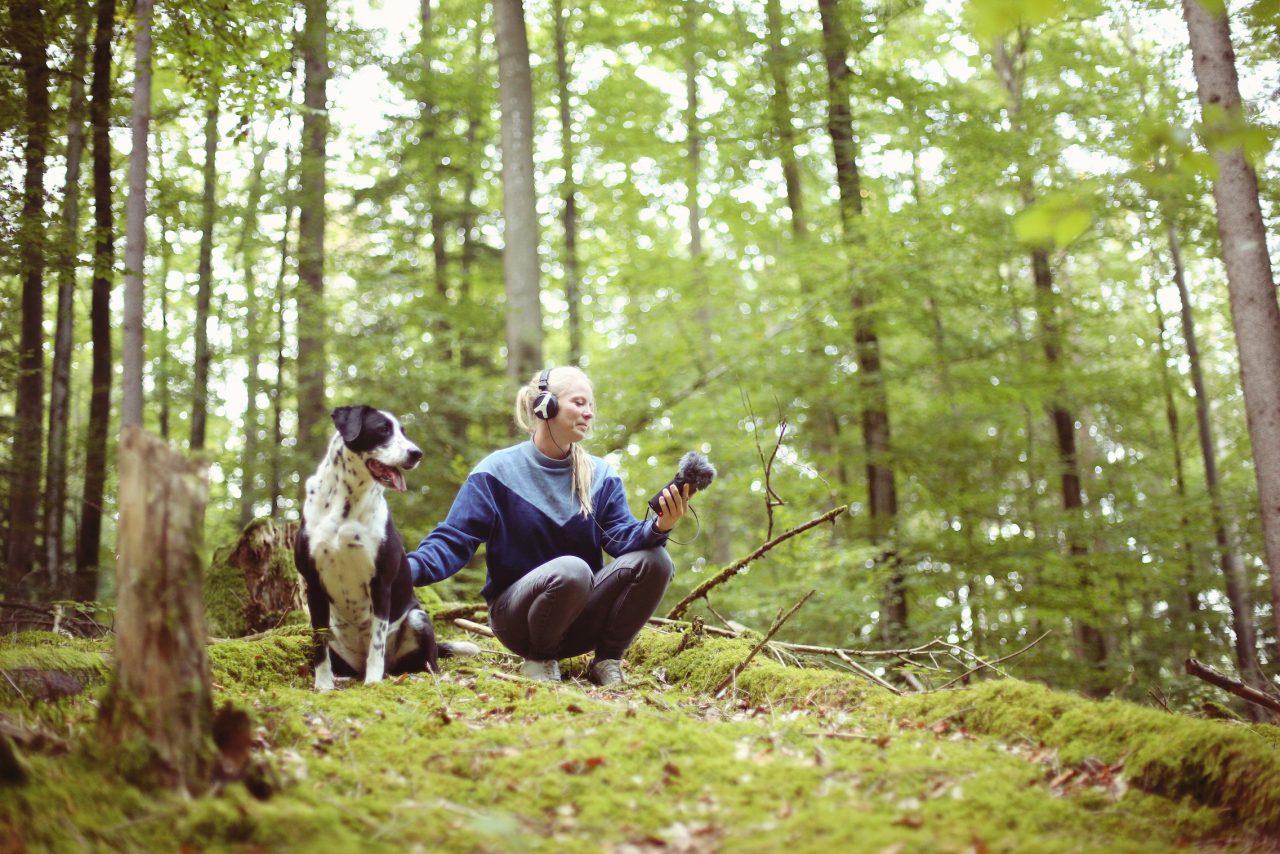 Frau (her tree) nimmt im Wald Geräusche auf. Neben ihr ein Hund.