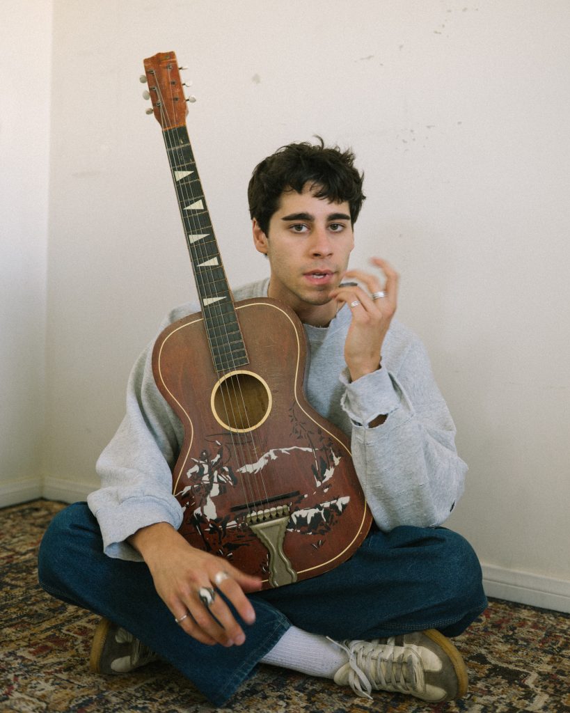 junge mit Gitarre, folk Musiker, natürlich, sitzt auf Teppichboden, schaut in die Kamera
